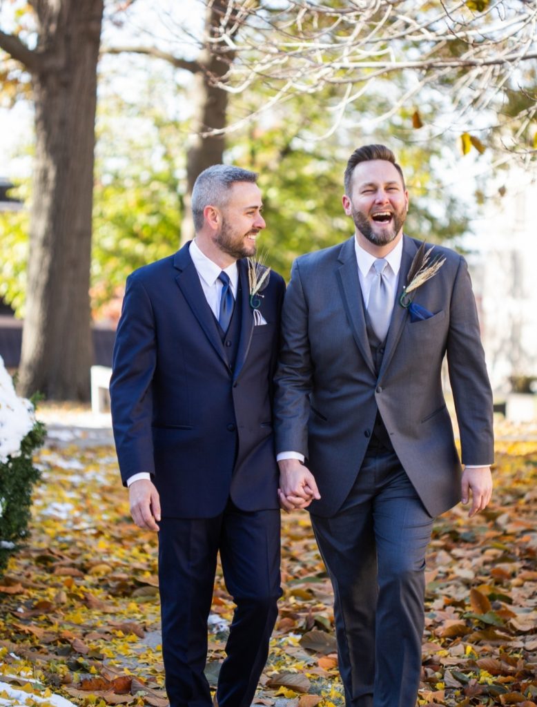Two men in suits laugh as they walk toward the camera holding hands